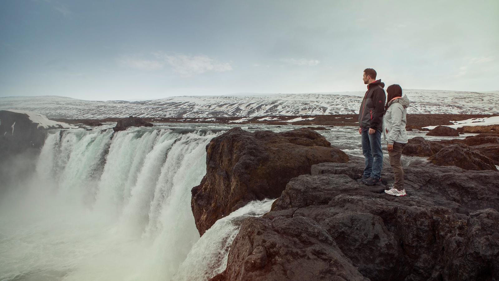 Two travellers standing on the edge of a clear blue waterfall in Iceland