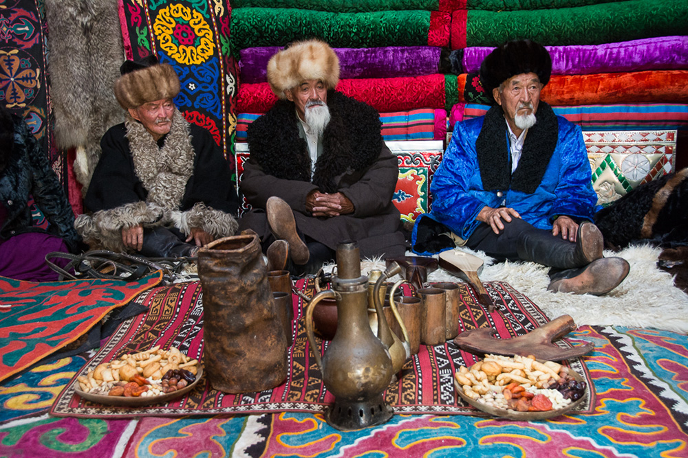 Kyrgyz elder men wait for guests in a yurt. Notice the colorful “shyrdak” carpets on the ground.